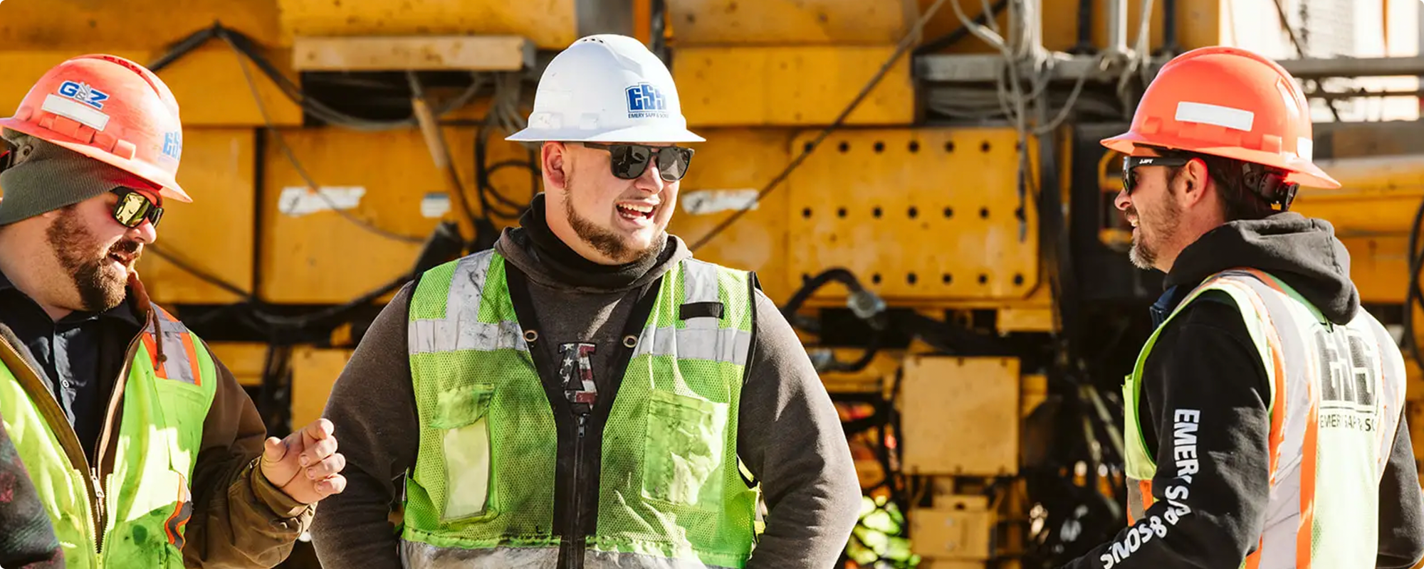 Three workers in safety gear talk at a site with yellow equipment.