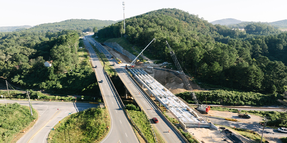 Highway bridge construction with cranes amid green hills, sunny day.