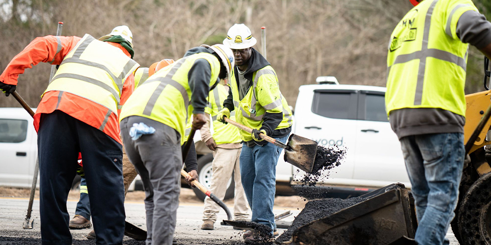 Workers spread asphalt on road; vehicles parked behind them.