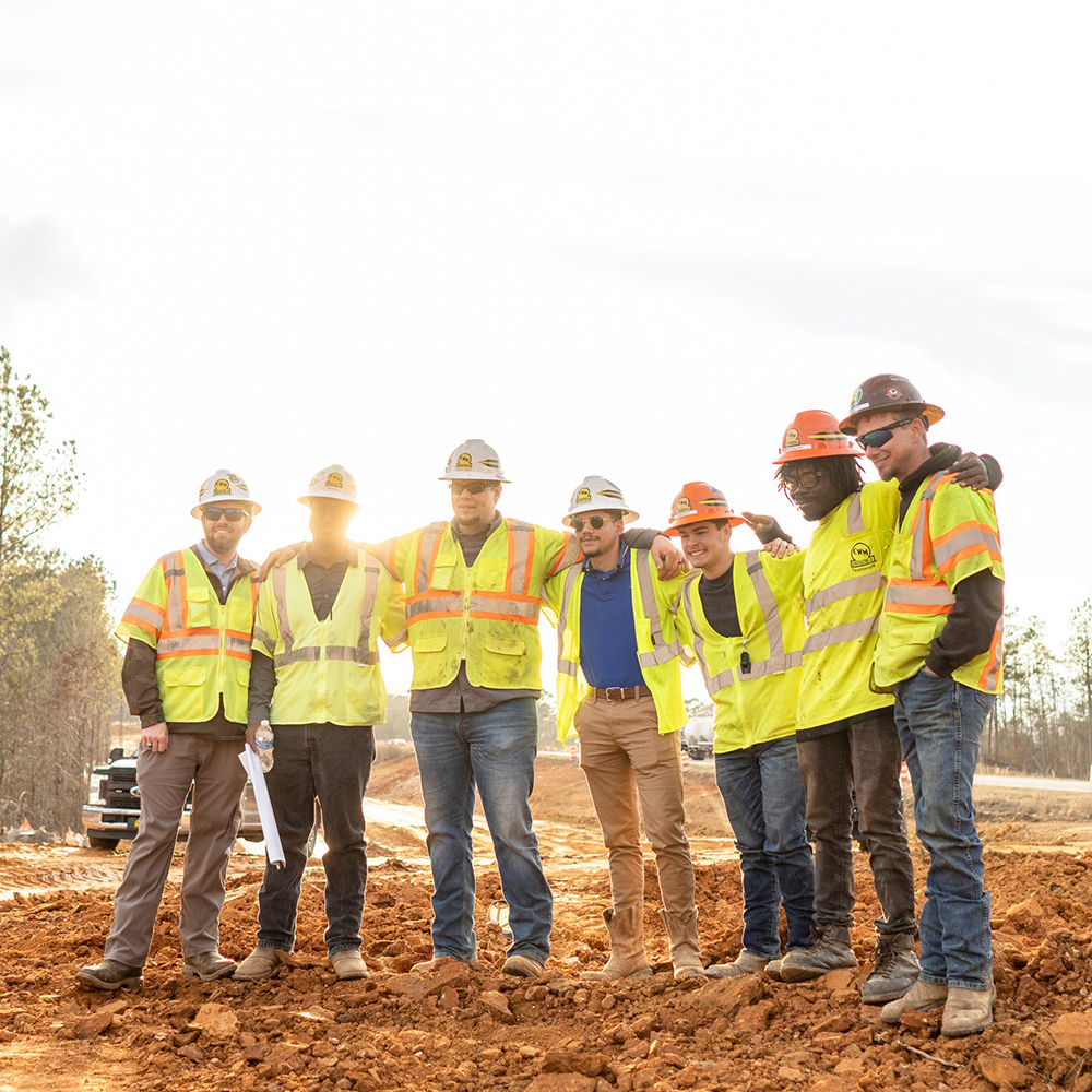 Six construction workers in vests and helmets pose on a dirt site.