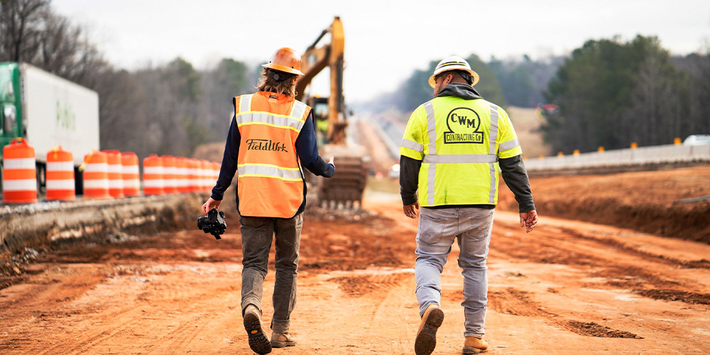 Two workers walk beside traffic barrels and excavator.
