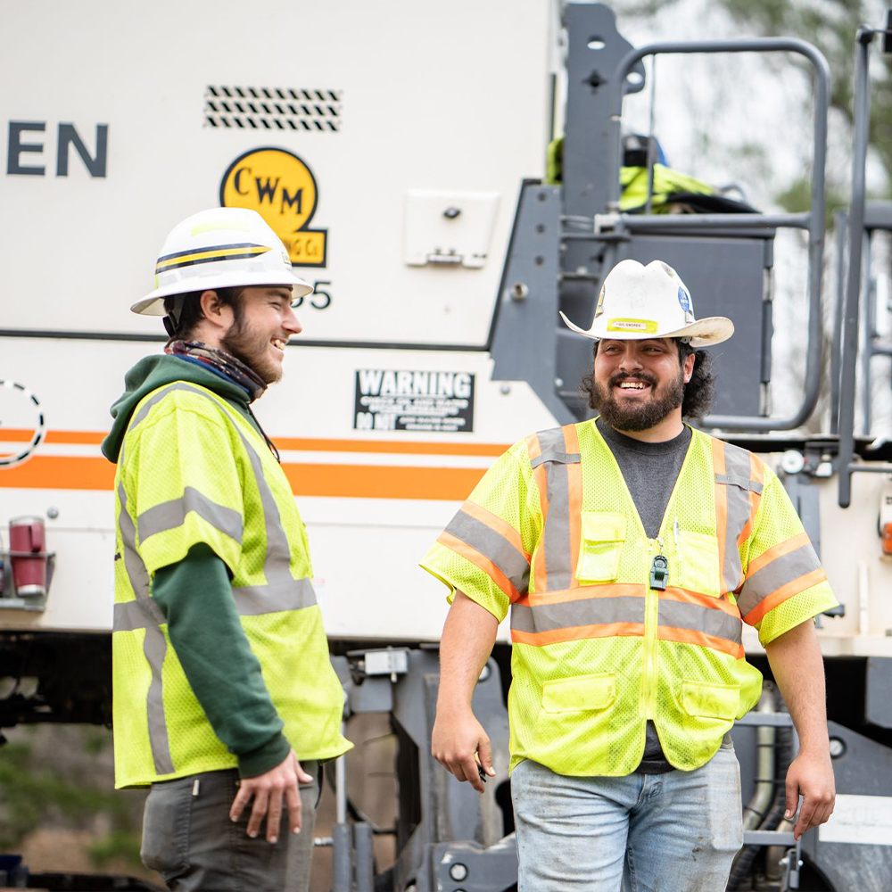 Two construction workers talk by industrial equipment outdoors.