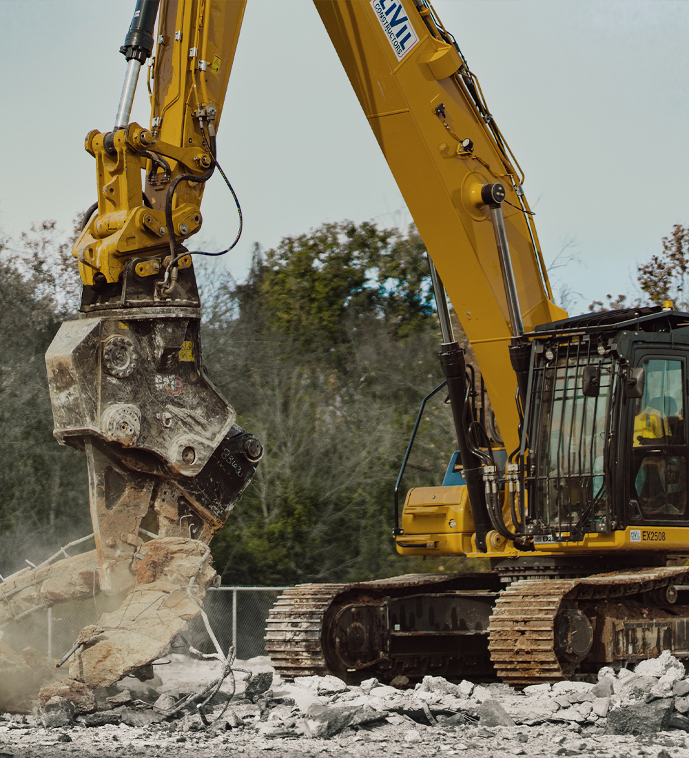 Yellow excavator breaking concrete with hydraulic attachment.