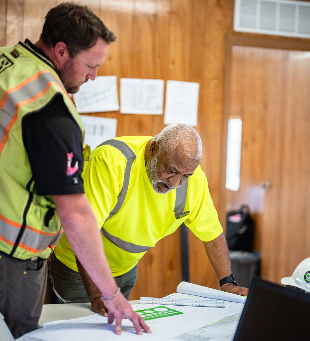 Two men in safety gear review blueprints in a wood-paneled room.