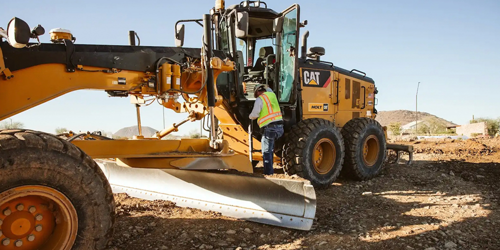 Worker in safety vest climbs into yellow CAT grader at site.