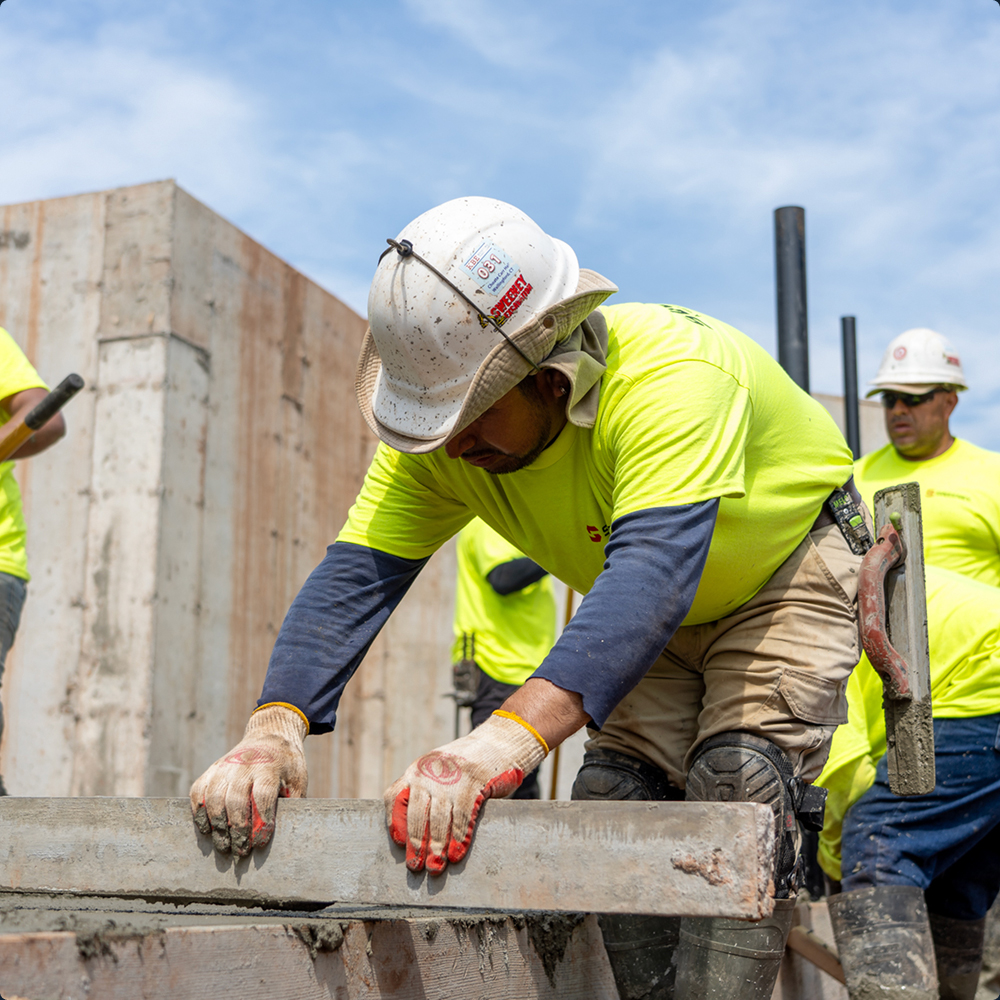Worker in yellow shirt levels concrete as others work nearby.