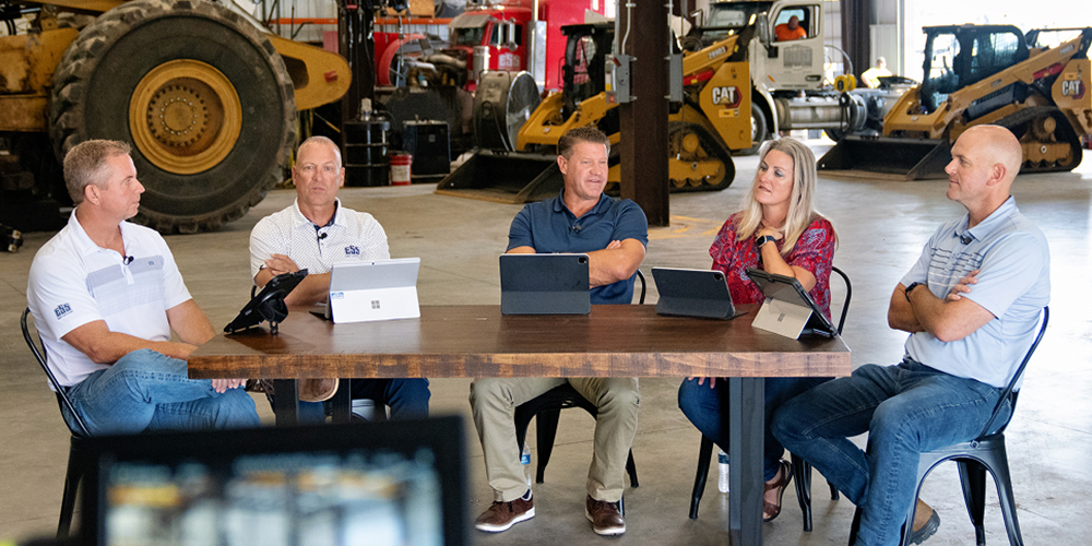 Five people at table with papers, tablets, and vehicles behind.