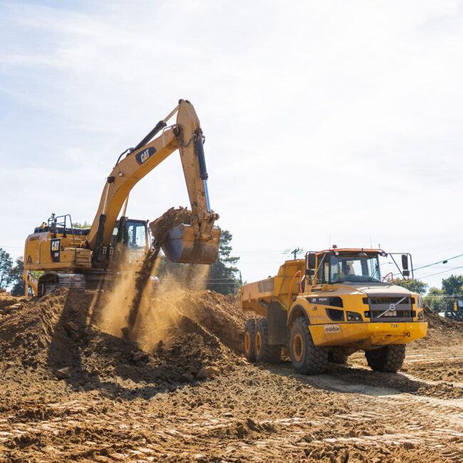 Yellow excavator loads dirt into yellow dump truck outdoors.