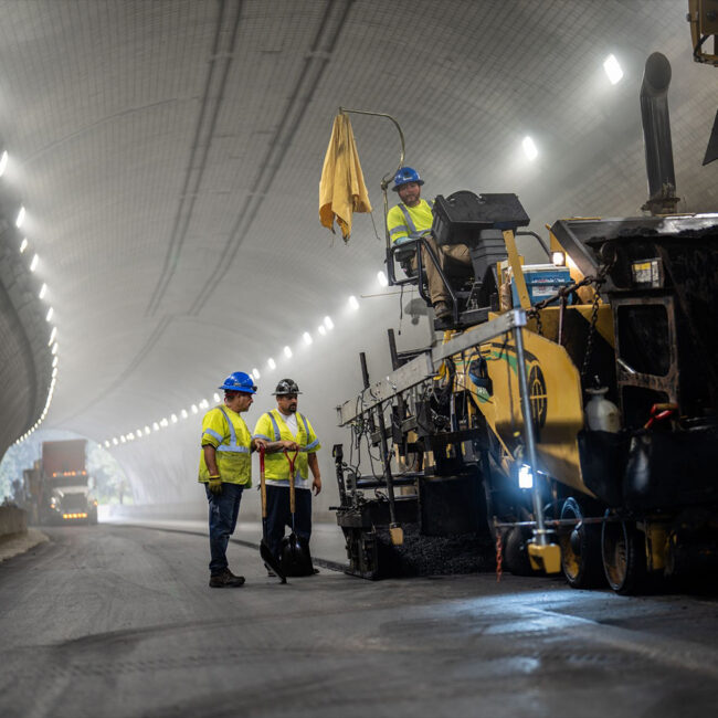 Construction workers in safety gear operate paving machinery in tunnel.