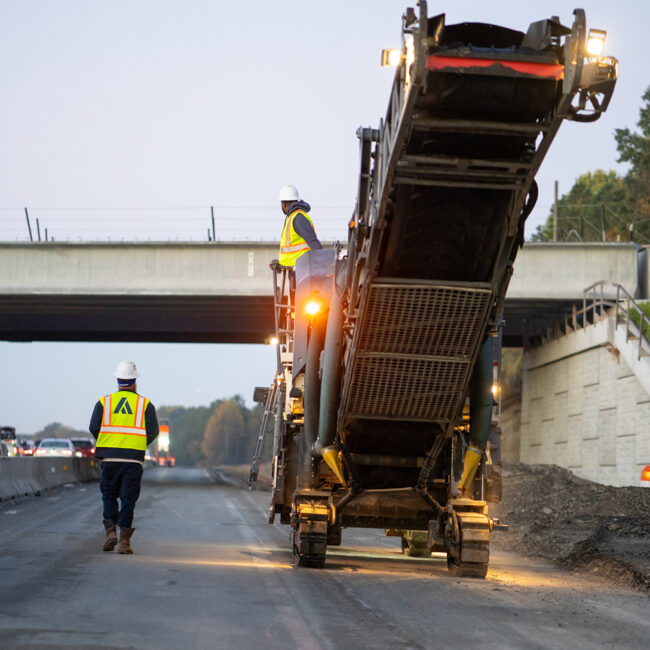Two workers in vests and helmets operate machinery by an overpass.