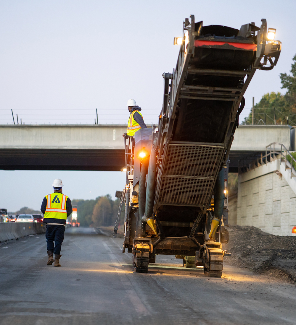 Two workers in vests and helmets operate machinery by an overpass.