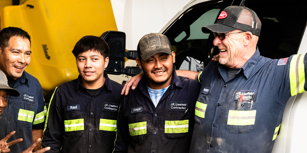 Four men in matching uniforms smile by a yellow truck.