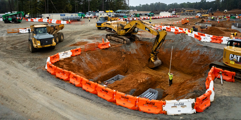Excavators dig pit, worker with measuring rod, orange barriers.