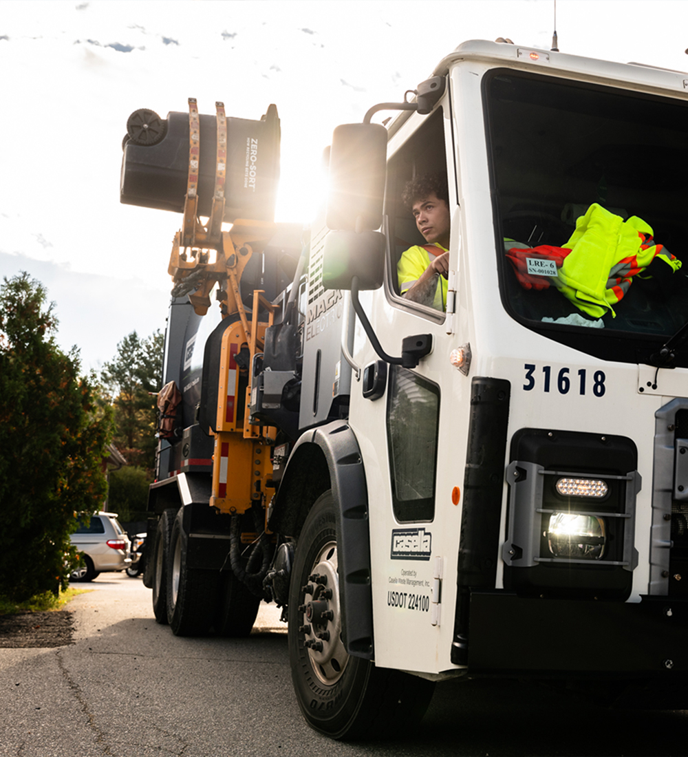 Worker drives white garbage truck with arm on sunny suburban street.