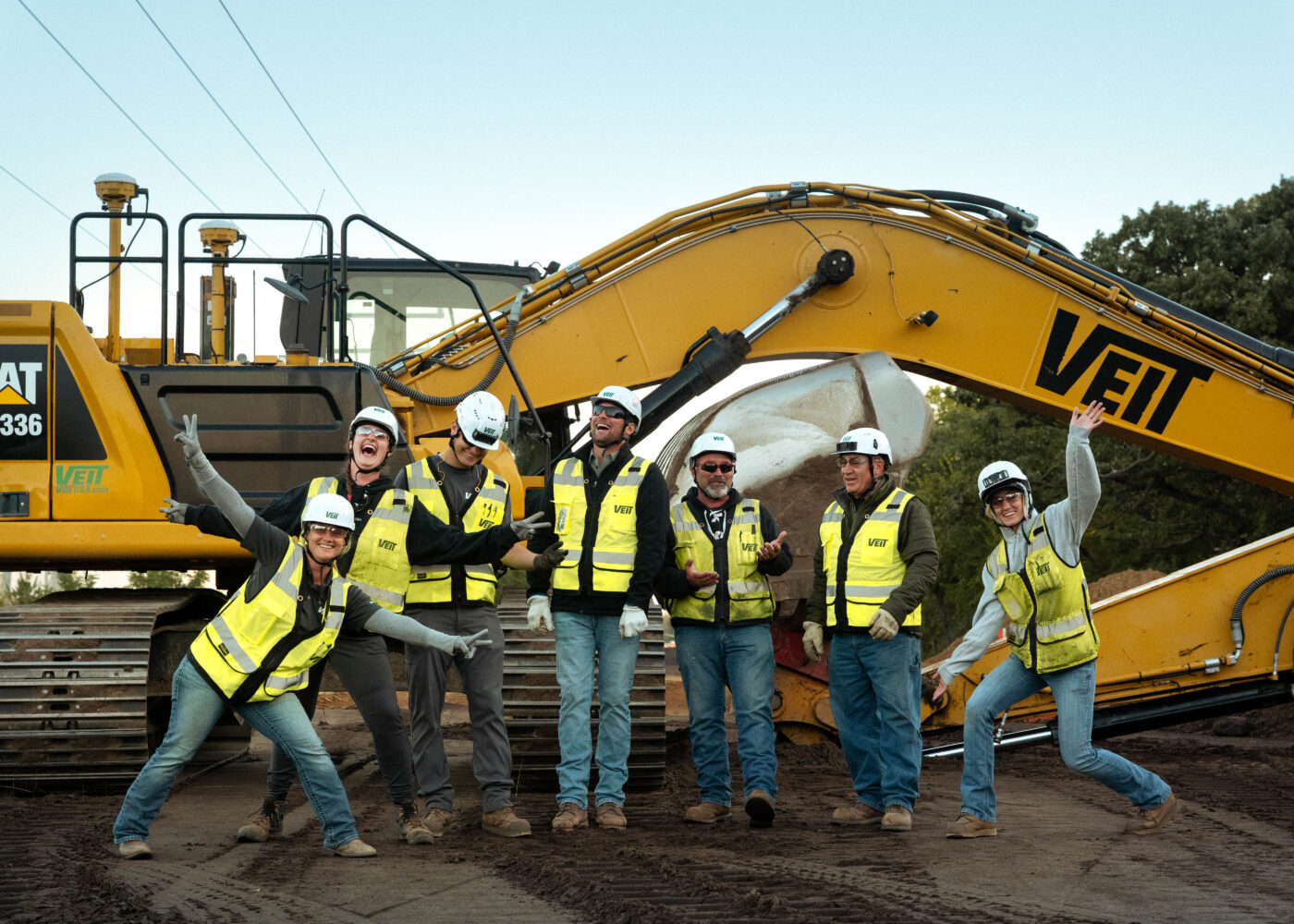 A work crew poses happily in front of a yellow excavator.