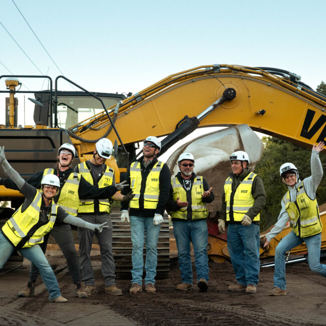 A work crew poses happily in front of a yellow excavator.