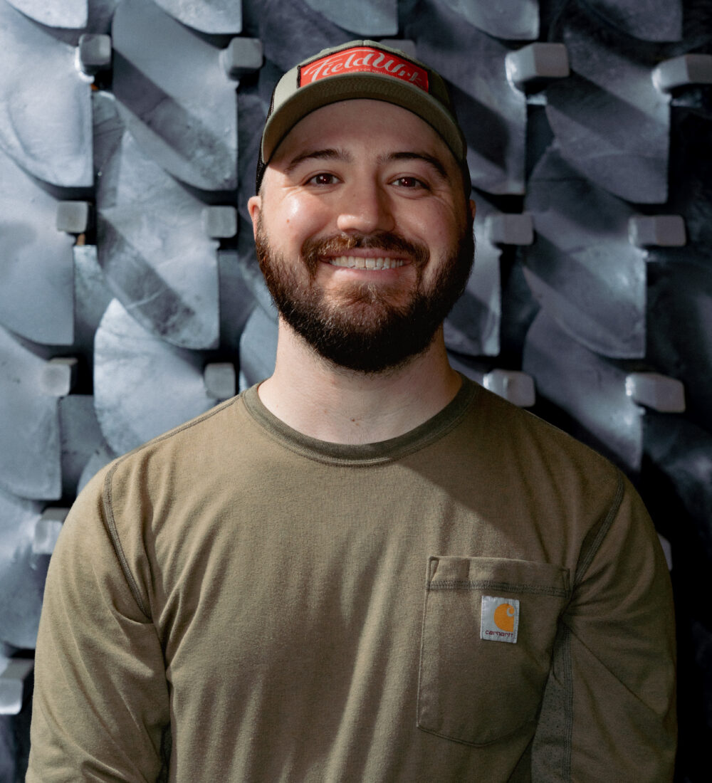 A man in a green shirt and a hat smiles at the camera in front of a gray background.