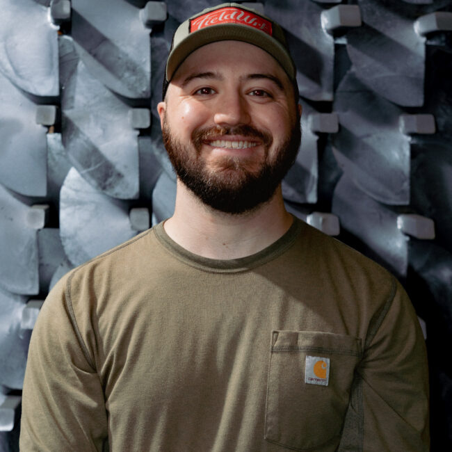A man in a green shirt and a hat smiles at the camera in front of a gray background.
